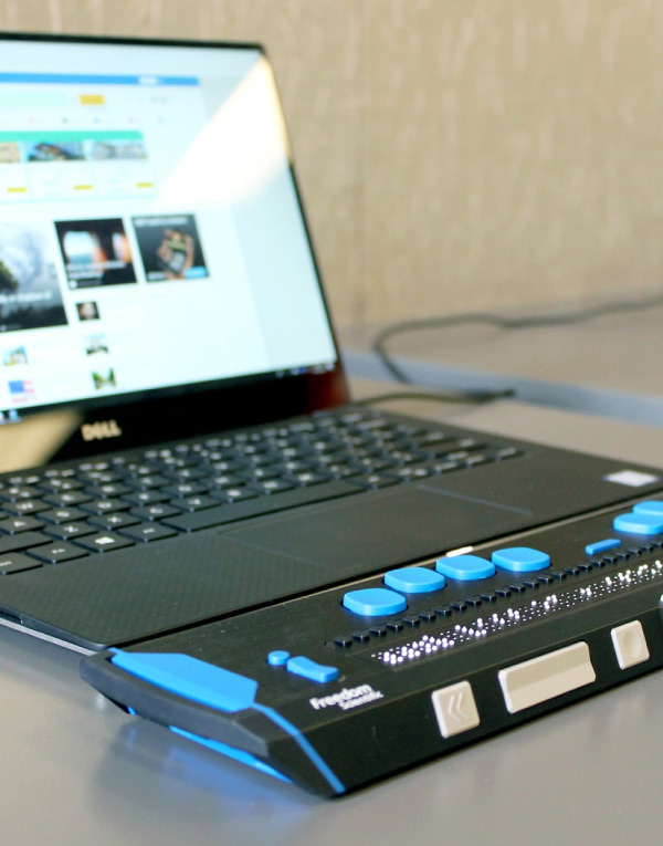 Auf dem Bild ist ein Laptop mit angeschlossener Braillezeile zu sehen. Im Vordergrund ist eine schwarze Braillezeile mit blauen Tasten und erhabenen Braillepunkten zu sehen, die als Hilfsmittel für blinde oder sehbehinderte Menschen dient. Im Hintergrund steht ein aufgeklappter Dell-Laptop mit geöffneter Internetseite auf dem Bildschirm. Beide Geräte stehen auf einem Tisch vor einer hellen Wand.